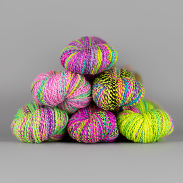 Skeins of Spincycle Dyed in the Wool sport weight yarn in a neon green, pink, and purple color gradient against a gray background.