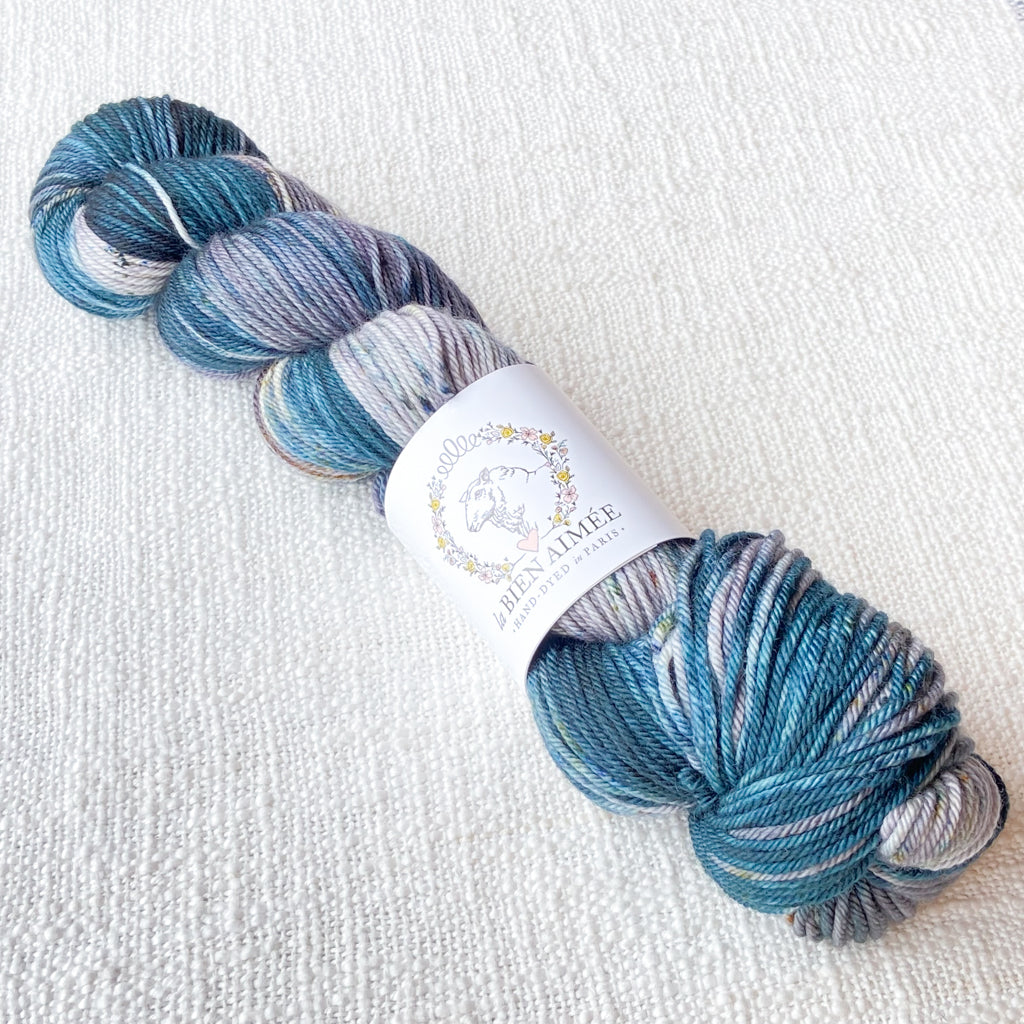 An image of a skein of DK weight yarn in shades of ocean blue, gray, and white with a white ‘La Bien Aimee’ label against a canvas background.