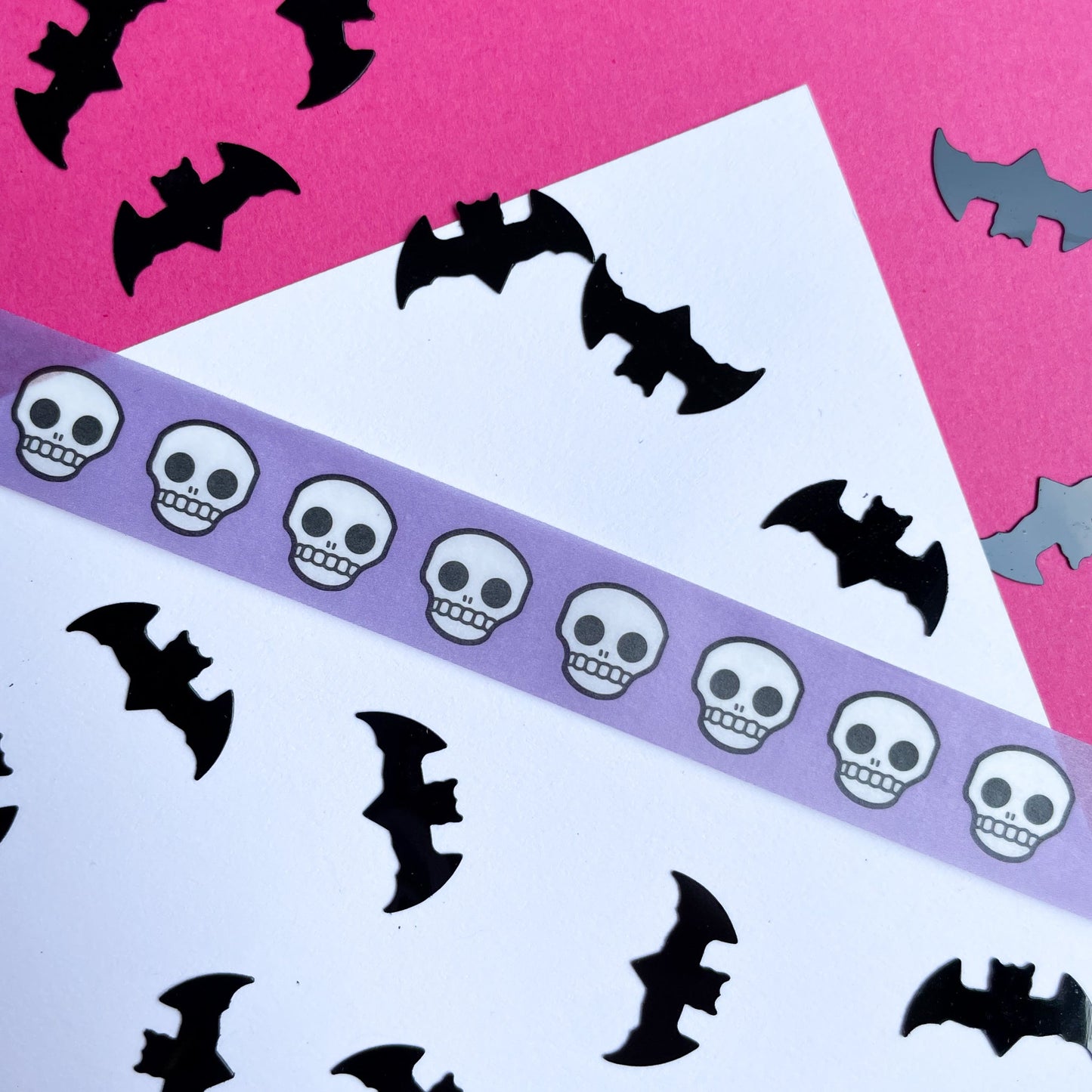 The corner of a piece of paper on a hot pink background. The paper has a piece of periwinkle washi tape with skulls on it stuck to it and bat confetti strewn about. 